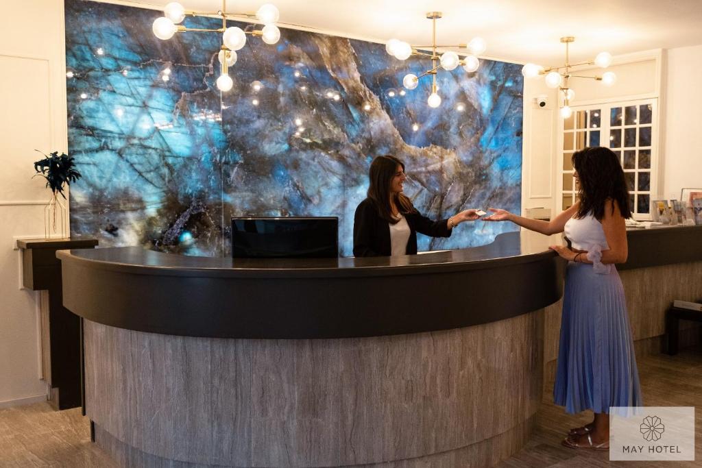 Recepcija modernā viesnīcā ar elegantu apgaismojumu un akmens fona sienu, kur viesmīlis pasniedz atslēgas viesim.