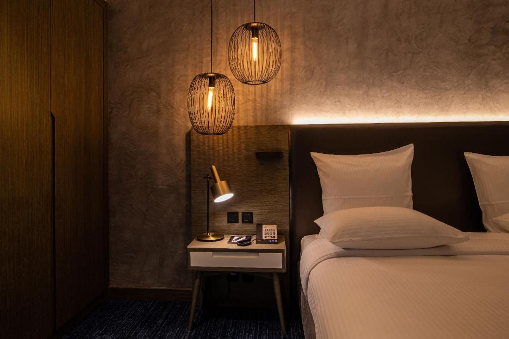 Mūsdienīgs guļamistabas interjers ar elegantiem gaismas ķermeņiem, modernu gultu un balto naktsgaldiņu, kas papildina siltu un viesmīlīgu atmosfēru.