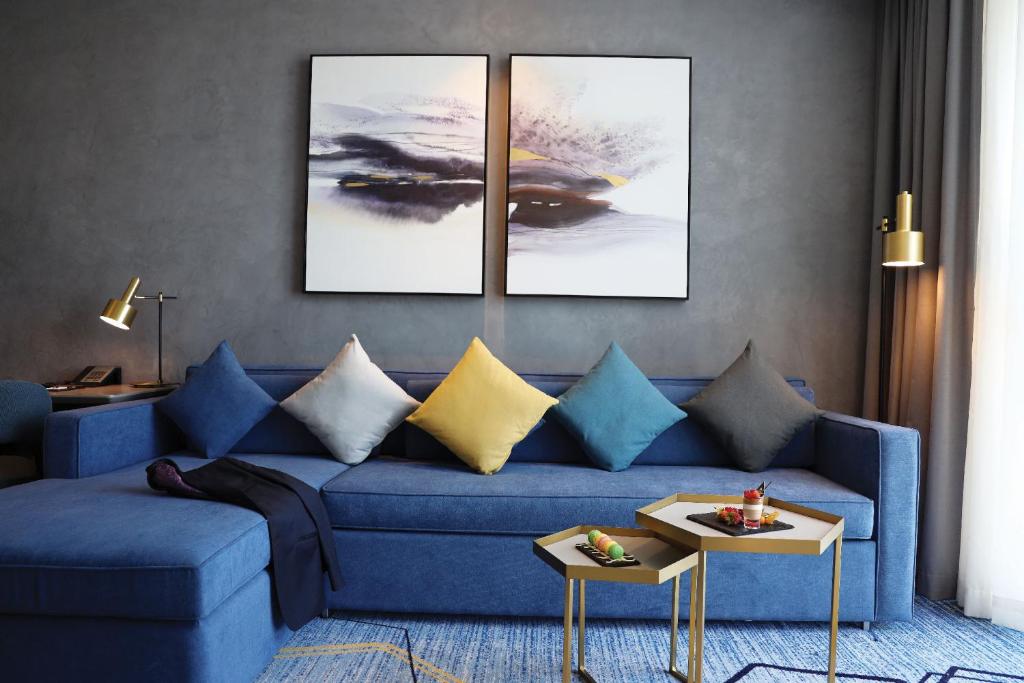 Mūsdienīgs viesistabas interjers ar zilu dīvānu, dekorēts ar spilveniem un divām mākslas gleznām uz sienas, kā arī zelta krāsas galdiņu ar uzkodām.