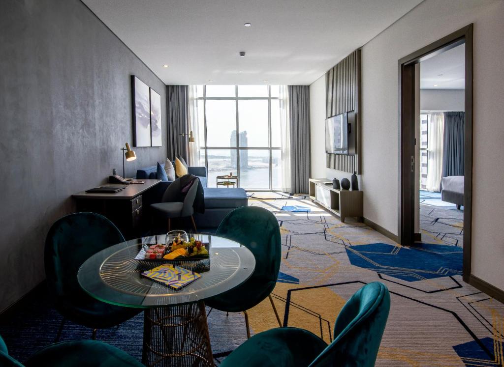 Mūsdienīgs viesnīcas numurs ar plašu dzīvojamo telpu, elegantām mēbelēm un skatu uz pilsētu. Uz galda ir augļu plates un žurnāli, radot komfortablu atmosfēru.