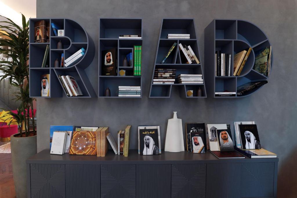 Grāmatu plaukts, kas veidots burtiem "READ", satur dažādas grāmatas un dekoratīvus priekšmetus, radot mūsdienīgu un radošu atmosfēru. Apakšā redzami arī citi izdevumi un estētiski elementi, kas papildina telpu.