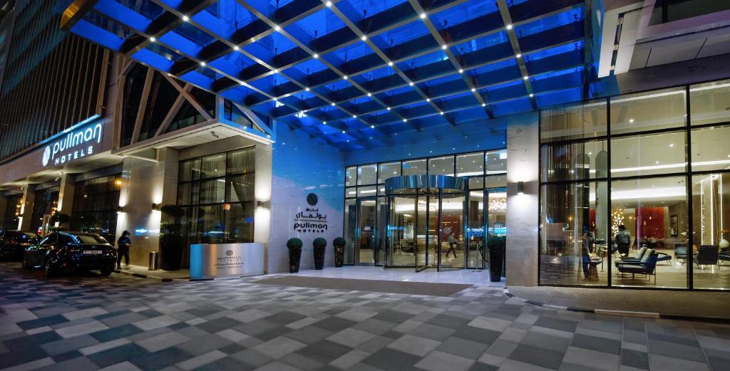 Mūsdienīga viesnīcas Pulman ieejas zona ar elegantu arhitektūru un apgaismojumu naktī. Viesnīcas logi atklāj stilīgu interjeru un viesnīcas nosaukums ir redzams uz ēkas fasādes.