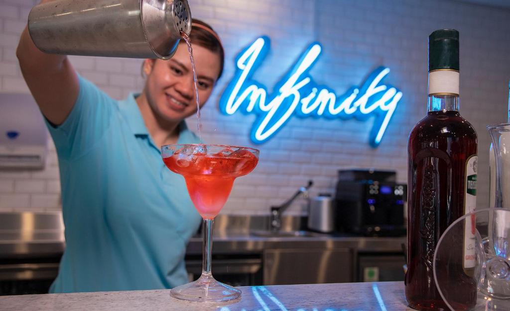 Bārmenis, kas ielej dzērienu glāzē, fonā ir neonā izgaismots "Infinity" uzraksts un dažādi alkoholiskie dzērieni.