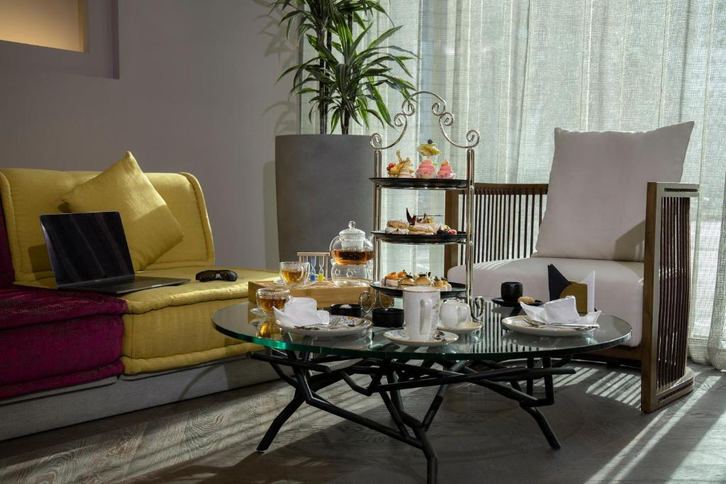 Mūsdienīgs viesistabas interjers ar dzeltenu un rozā dīvānu, stikla galdu un tējas komplektu, uz galda ir maizītes un saldumi. Aiz galda redzama liela logs ar gaismu un zaļu augu.