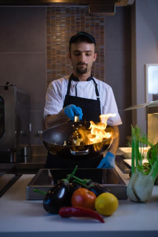 Šefpavārs gatavo ēdienu wok pannā, radot liesmas, kamēr priekšplānā ir dažādi dārzeņi, piemēram, baklažāni, paprika un citroni.