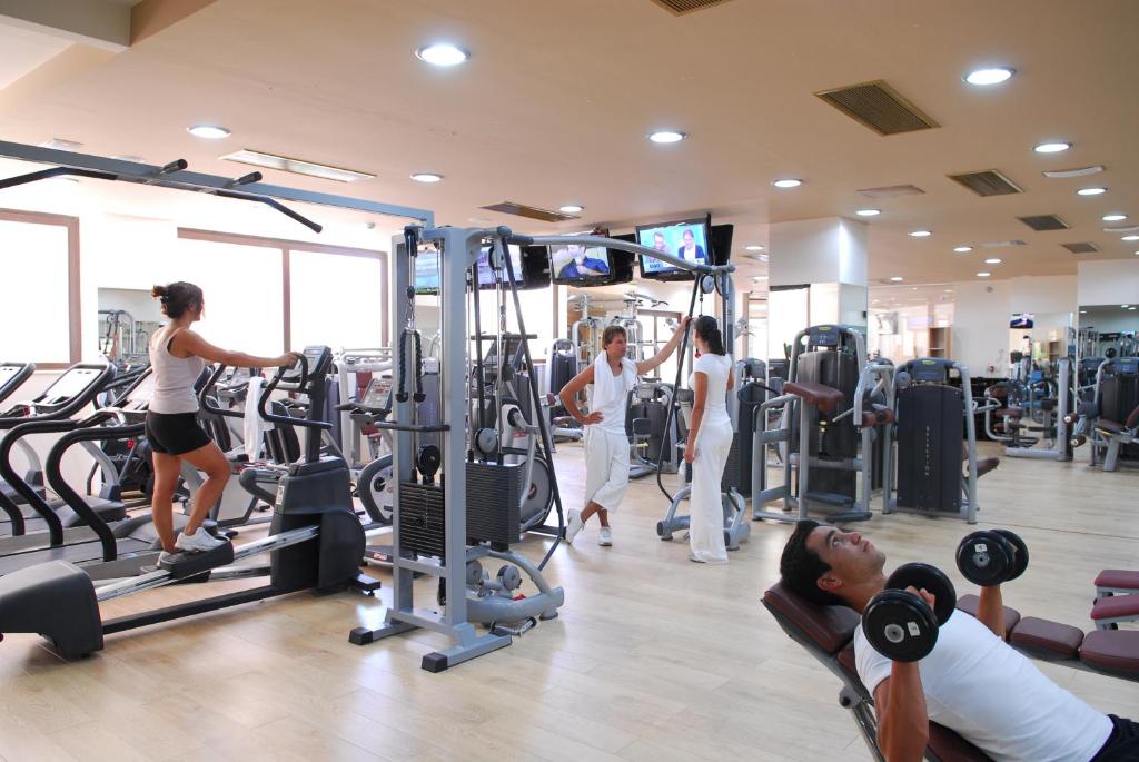 Mūsdienīga sporta zāle ar dažādām fitnesa iekārtām, kur cilvēki nodarbojas ar vingrošanu un spēka treniņiem. Gaisma ir spilgta, un ir redzamas vairākas sporta mašīnas.