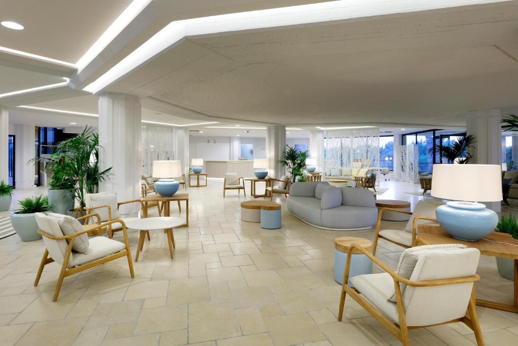 Mūsdienīgs viesnīcas vestibils ar gaišām krāsām, saplūstošām mēbelēm un zaļām augiem, radot relaksējošu atmosfēru. Apgaismojums un interjera dizains piešķir telpai modernu un viesmīlīgu noskaņu.