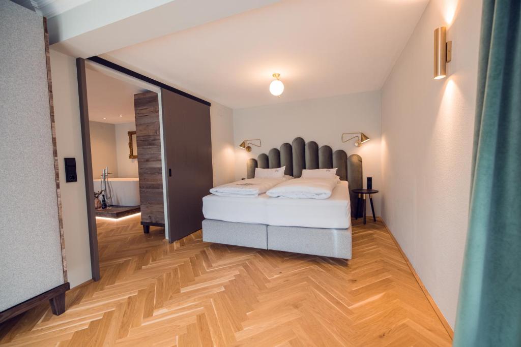 Mūsdienīgs guļamistabas interjers ar elegantu gultu un koka grīdu, kas rada siltu atmosfēru. Telpā ir arī slīdošās durvis un lampas, kas piešķir modernu akcentu.