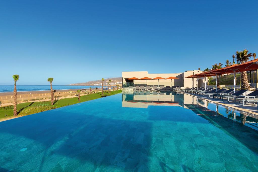 Elegants baseins ar skatu uz pludmali un zilo okeānu. Apkārt baseinam ir sauļošanās krēsli un palmām apstādīta teritorija, kas rada relaksējošu atmosfēru.