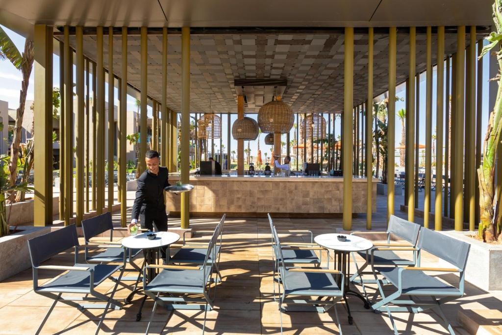 Mūsdienīgs restorāns ar atvērtu terasi, kur viesmīlis pasniedz dzērienus, un redzama stilīga interjera dizaina elementu kombinācija. Pamanāmi zaļumi un eleganti galdi ar krēsliem.