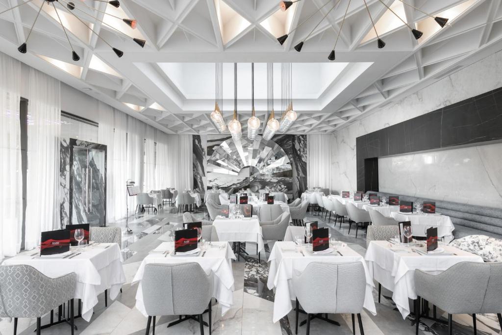 Mūsdienīgs restorāna interjers ar elegantiem galdiem, baltām galdautām un stilīgu apgaismojumu. Telpas dizainā apvienots mūsdienīgs un grezns stils, radot patīkamu atmosfēru.