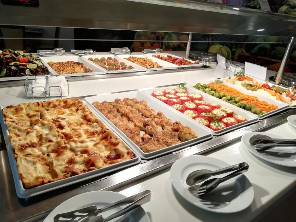 Dažādu ēdienu bārs ar lasi, vistas gaļu un dārzeņiem, kas sakārtoti uz baltiem traukiem. Iespēja izvēlēties no plaša ēdienu klāsta, tostarp salātiem un makaronu ēdieniem.