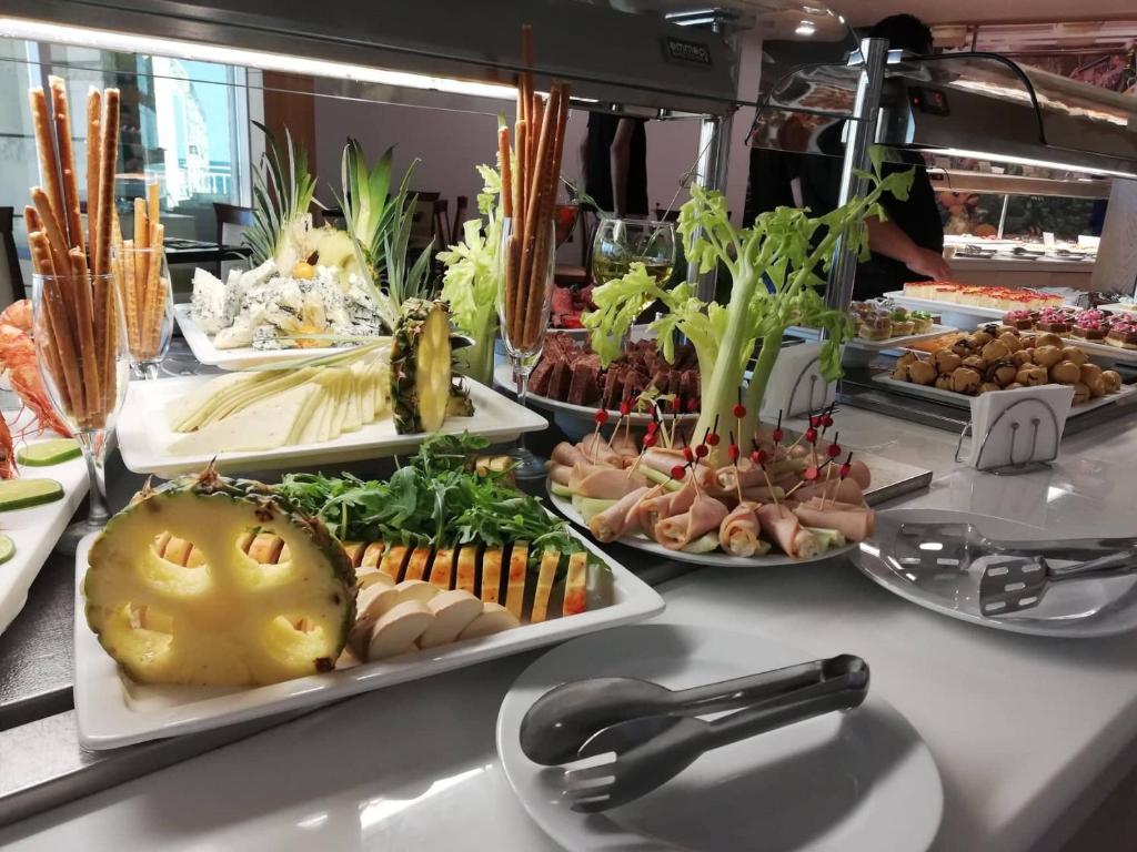 Bagātīga uzkodu bufete ar dažādām ēdienu izvēlēm, tostarp augļiem, dārzeņiem un gaļas izstrādājumiem. Uzkodas ir izkārtotas estētiski, radot vilinošu atmosfēru.