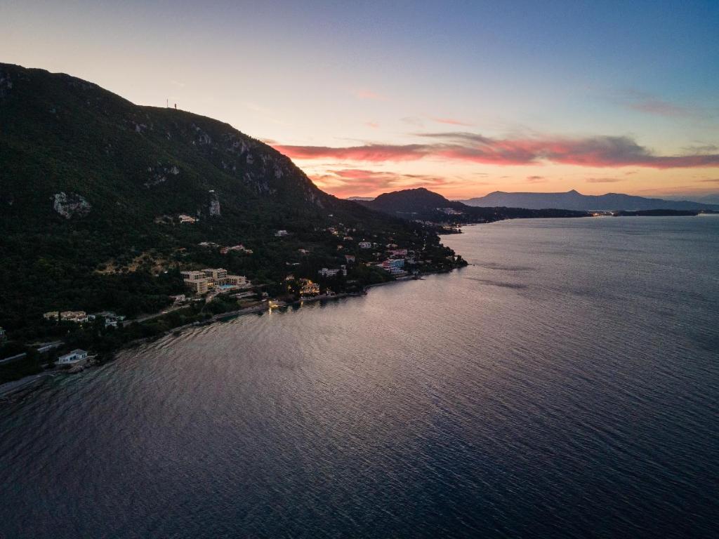 Krāsains saulriets virs kalnainas piekrastes, kur redzamas apbūves un jūras viļņi. Tumsā atspoguļojušās gaismas pie ūdens malas veido mierīgu un gleznainu ainu.