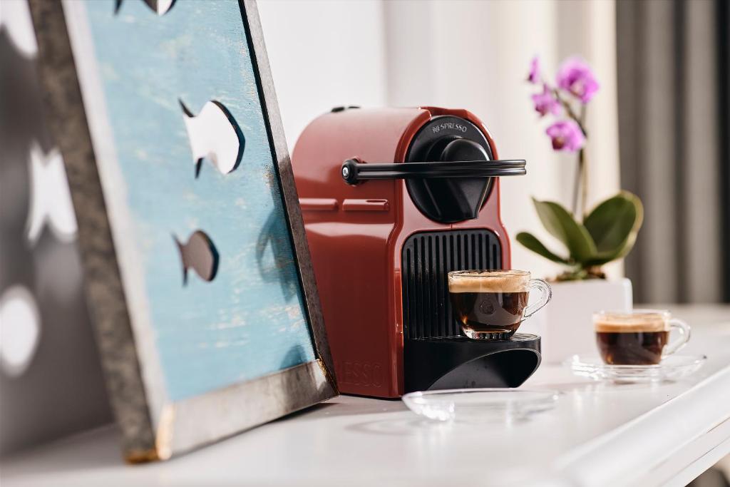 Sarkans kafijas aparāts ar divām kafijas krūzēm un ziedu fonā, uzsvērts moderns interjers.