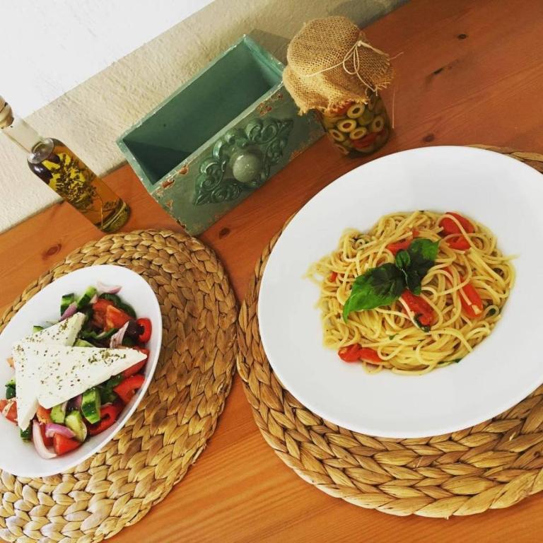 Garšīgs makaronu ēdiens ar tomātiem un baziliku, līdzās svaiga dārzeņu salāti ar fetas sieru. Uz galda ir arī olīveļļa un dekoratīva trauka ar olīvām.