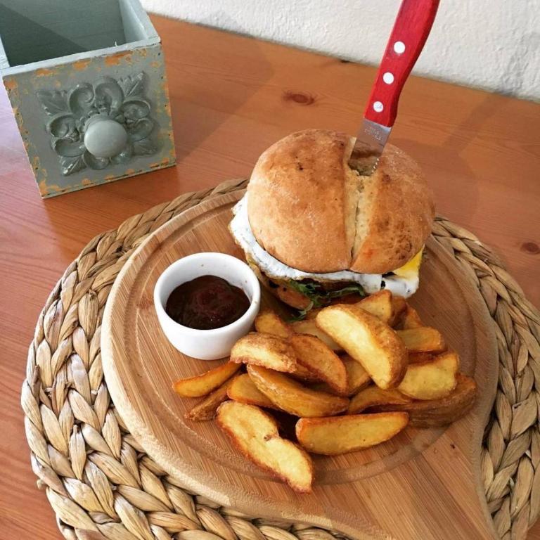 Svaigs burgeris ar olu un dārzeņiem, pasniegts uz koka šķīvja kopā ar ceptiem kartupeļiem un mērci.