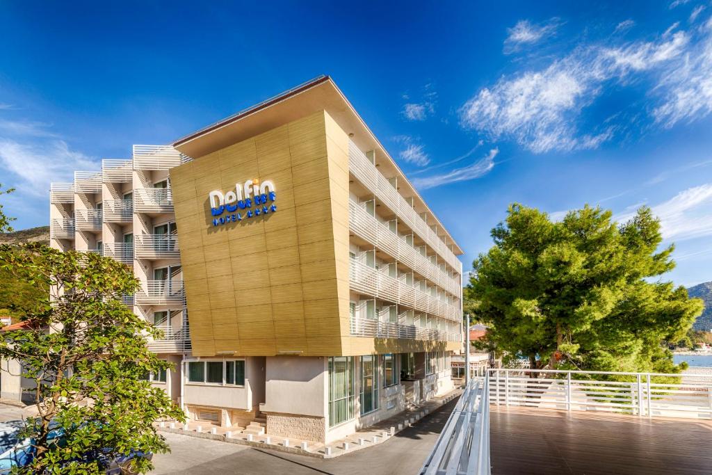 Delfin viesnīca ar modernu arhitektūru, kas atrodas blakus jūrai. Apkārtne ir zaļa, ar kokiem un skaidru debesu fonu.