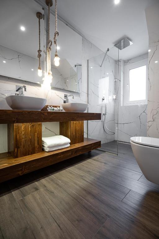 Mūsdienīgs vannas istabas interjers ar divām izlietnēm uz koka galda, akmens sienām un stikla dušu. Gaismas piekari un eleganta flīžu grīda piešķir telpai modernu izskatu.