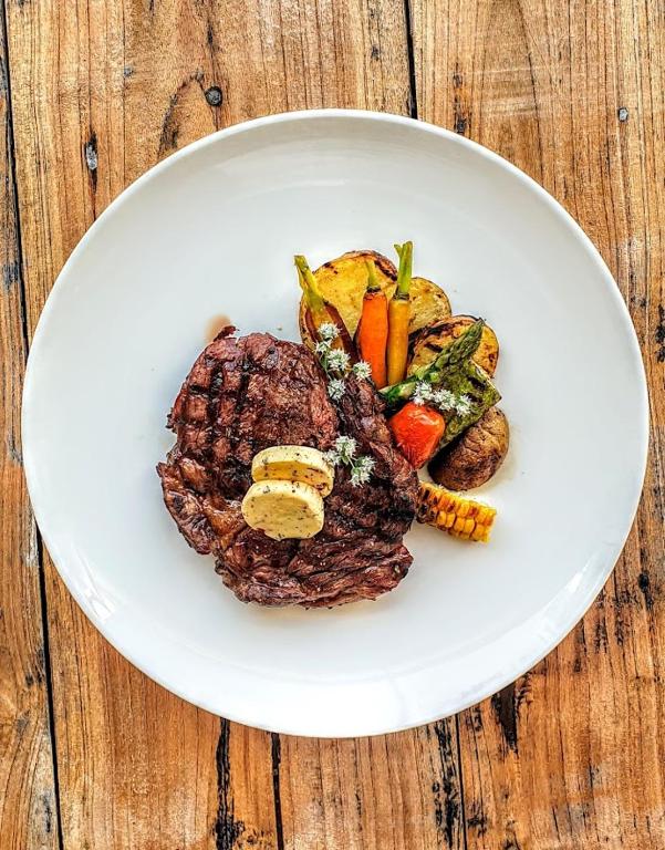 Grilēta gaļa ar dārzeņiem un sviesta gabaliņiem uz baltas šķīvja, kas novietots uz koka galda.