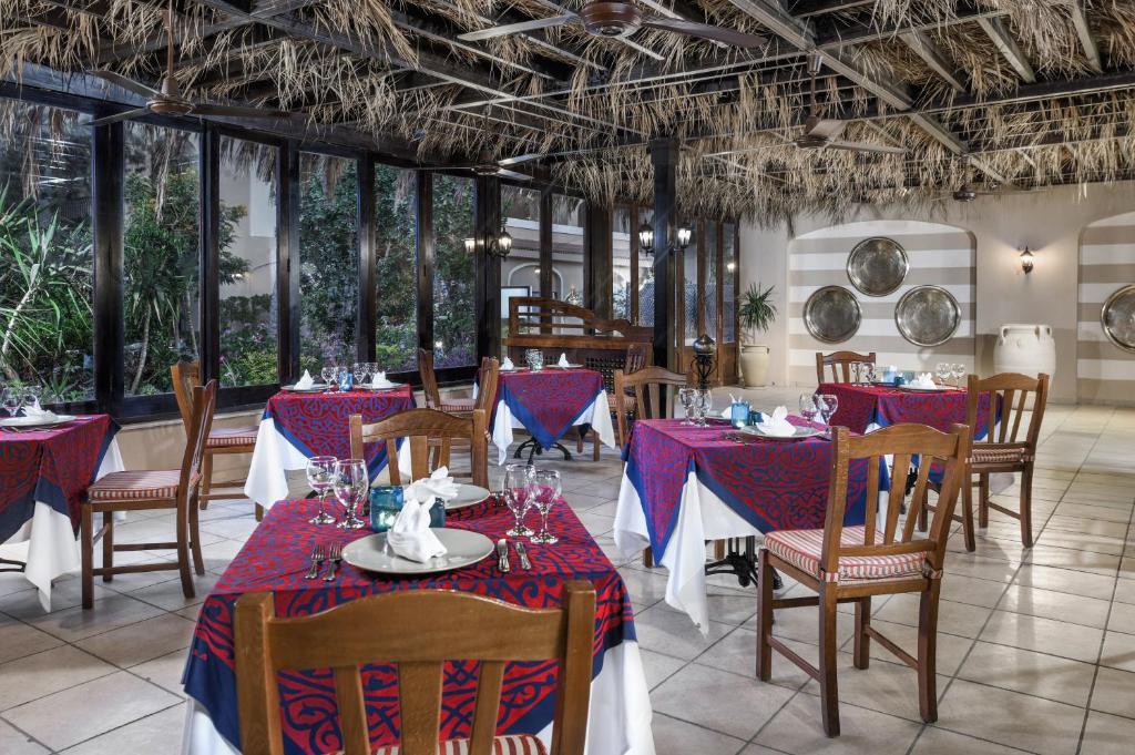 Elegants restorāns ar sarkaniem un ziliem galdiem, kas rotāti ar baltiem galdautiem. Telpa ar dabīgu apgaismojumu un tropiskiem augiem rada patīkamu atmosfēru.
