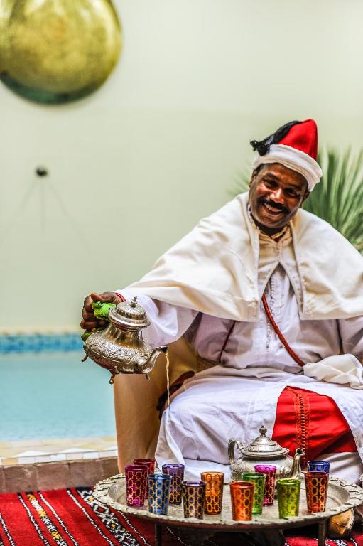 Vīrietis tradicionālā apģērbā pasniedz tēju no sudraba tējas kanna, sēžot pie galda ar krāsainām tējas krūzītēm. Fons ietver peldbaseinu un dekoratīvus elementus.