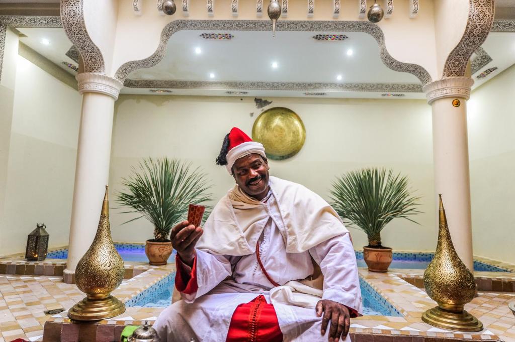 Vīrietis tradicionālā apģērbā ar sarkanu cepuri sēž dekoratīvā telpā ar palmām un zeltītām lampām. Fons ir rotāts ar arābu arhitektūras elementiem, radot relaksējošu atmosfēru.