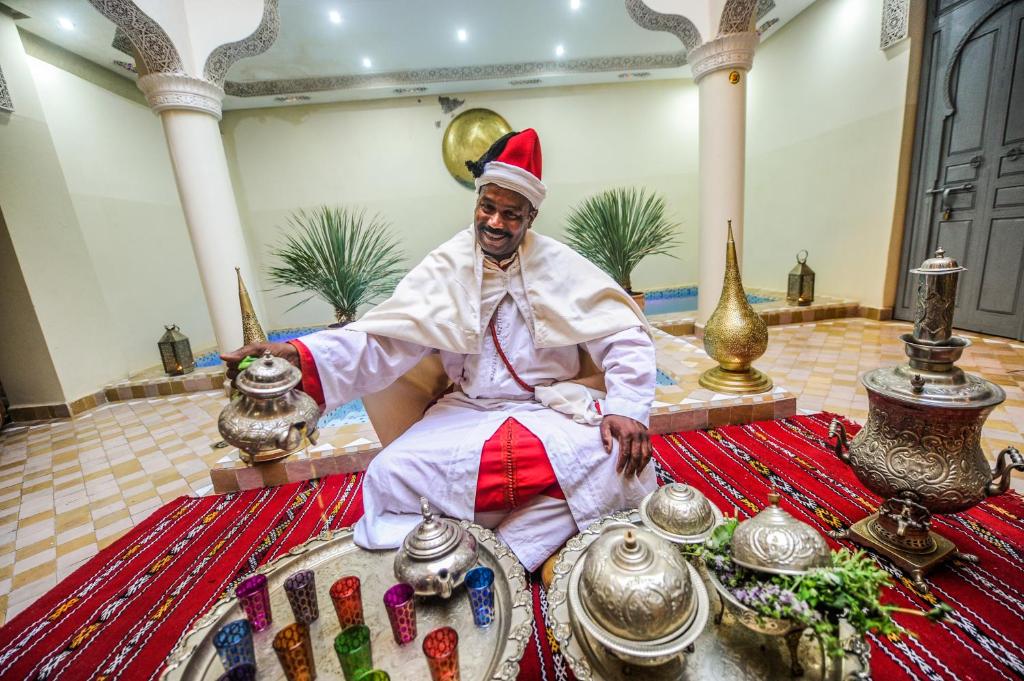 Vīrietis tradicionālajā apģērbā pasniedz tēju, sēžot uz grīdas tradicionālajā marokāņu interjerā. Apkārt redzami sudraba trauki un krāsaini stikla glāzes, radot autentisku atmosfēru.