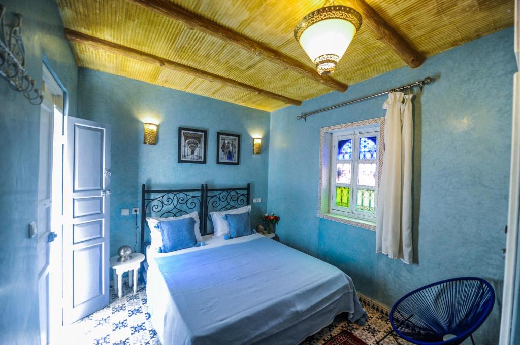 Mūsdienīga guļamistaba ar zilām sienām, dekoratīvām lampām un rakstainu grīdu. Gulta ar baltu gultasveļu un spilveniem, blakus neliels galdiņš un krēsls. Logā krāsaini stikli, radot mājīgu atmosfēru.