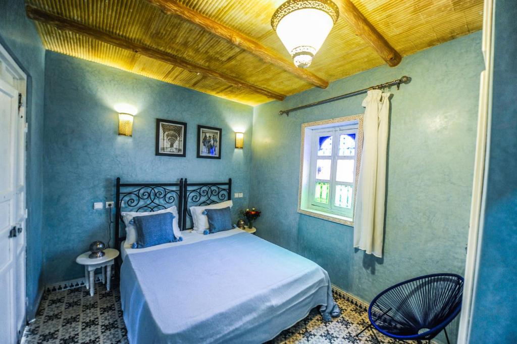 Mūsdienīga guļamistaba ar zilu sienu un koka griestiem, kurā ir divguļamā gulta, dekoratīvas lampas un krāsains logs. Atmosfēra ir mājīga un aicinoša, piedāvājot relaksējošu vidi.