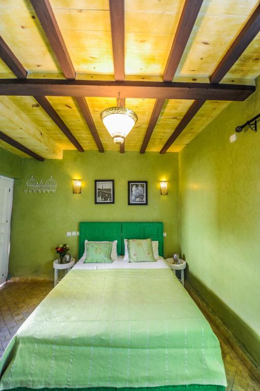 Mūsdienīgs guļamistabas interjers ar zaļu gultu un koka sijām griestos, radot siltu un viesmīlīgu atmosfēru. Sienas rotā mākslas darbi un siltas apgaismojuma lampas.