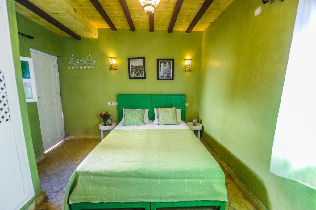 Mūsdienīgs guļamistabas interjers ar zaļu sienu apdari, koka griestiem un ērtu gultu. Uz sienām ir dekoratīvi attēli, un telpu izgaismo siltas gaismas avoti.