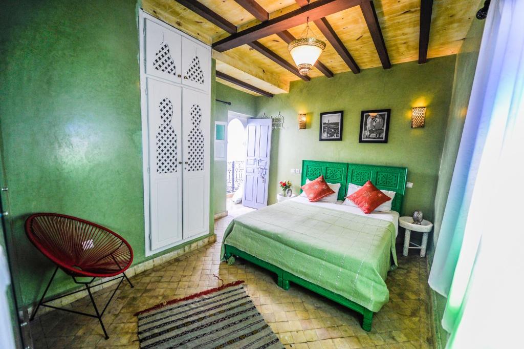 Mūsdienīgs guļamistabas interjers ar zaļām sienām, divguļamo gultu ar spilveniem un stilīgu krēslu. Telpā ir pieejami gaismas avoti un dekoratīvas fotogrāfijas uz sienām.