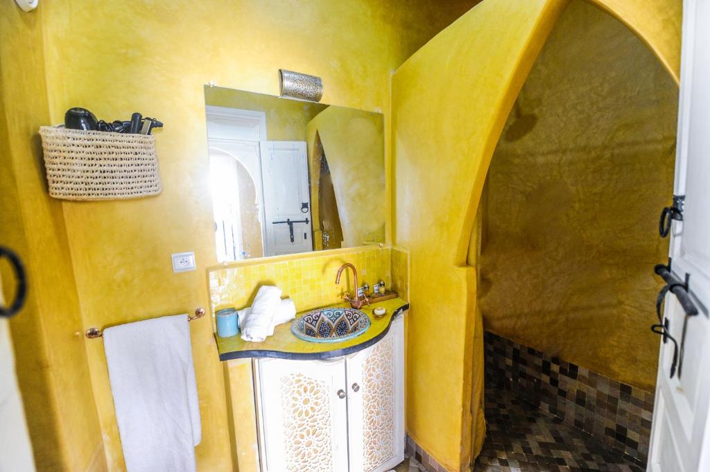 Mūsdienīga vannas istaba ar spilgtām dzeltenām sienām, tradicionālu izskatu un elegantu izlietni ar mozaīkas detaļām. Telpā ir iekļauta dušas zona un stilīgs spogulis.