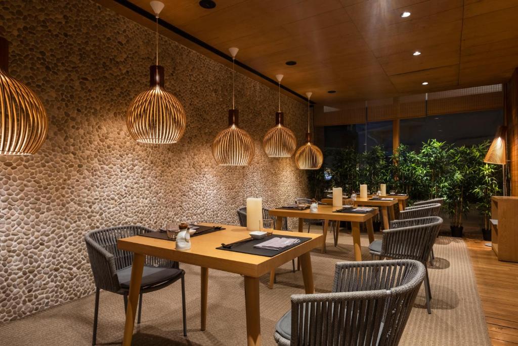 Mūsdienīgs restorāns ar dabīgiem akmeņiem sienā un elegantiem koka galdiem, apgaismots ar stilīgām lampām, radot siltu un aicinošu atmosfēru.