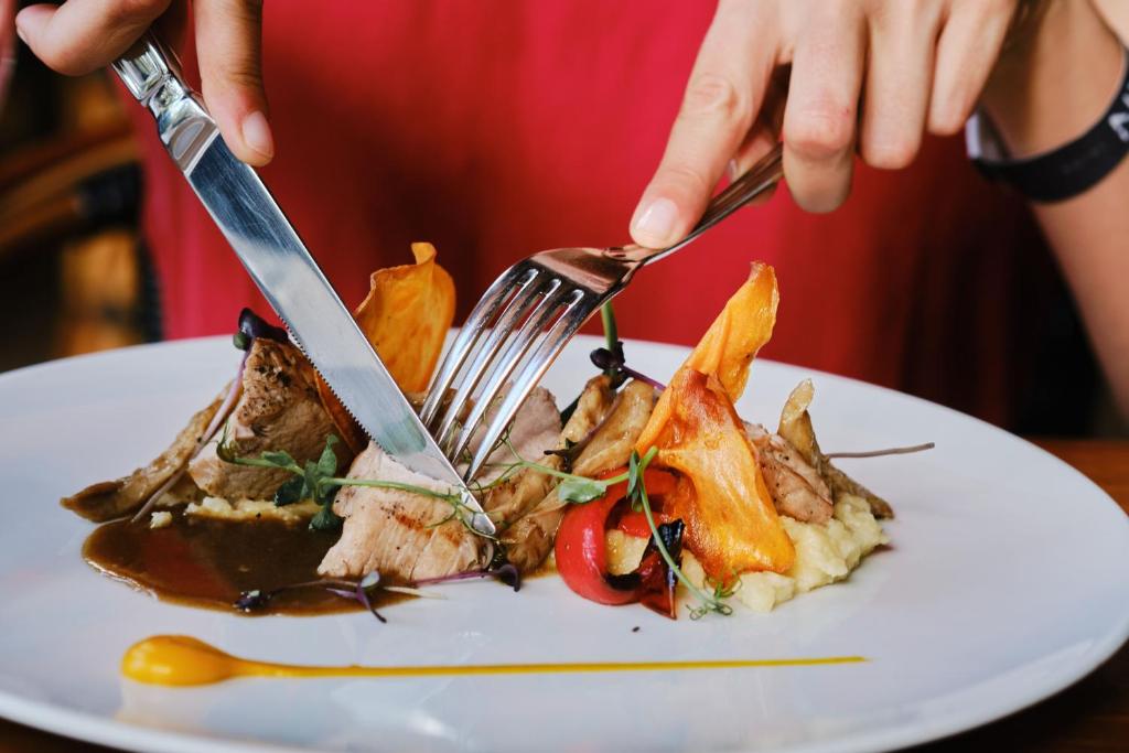 Skaisti pasniegts ēdiens ar gaļu, dārzeņiem un mērci, ko kāds griež ar nazi un dakšiņu. Plāksne ir dekorēta ar krāsainiem elementiem, radot apetīti rosinošu izskatu.