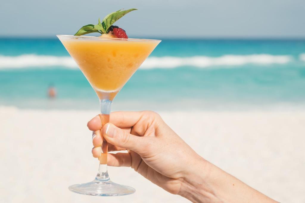 Gatavs kokteilis ar augļu dekorāciju tiek turēts rokā, pretī pludmalei ar smilšu krastu un jūras viļņiem fonā.