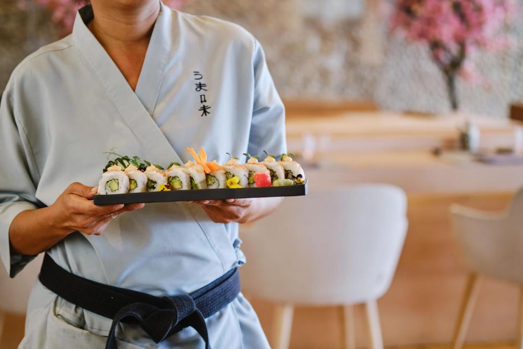 Šefpavārs tērptā tradicionālā apģērbā tur sushī šķīvi ar dažādām garšīgām rullīšiem, kas rotāti ar dārzeņiem un ziediem, restorāna interjerā ar rozā ziediem fonā.