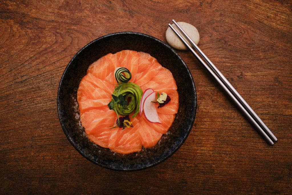 Svaigi griezta laša porcija ar dārzeņu dekorācijām, pasniegta melnā traukā uz koka virsmas, līdzās ir ēdamkarote un nūjiņas.