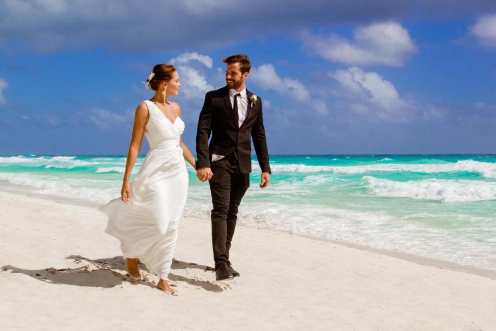 Jaunlaulātie pastaigā pa pludmali, baudot mierīgu atmosfēru un skaistu skatu uz jūru. Viņu apģērbs ir elegants un piemērots kāzu svinībām.