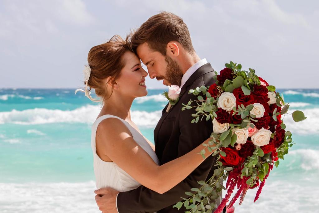 Jaunlaulātie pāris, kas laimīgi skskata viens uz otru pludmalē, turot rokās krāšņu ziedu pušķi. Fons ir skaists jūras skats ar viļņiem.
