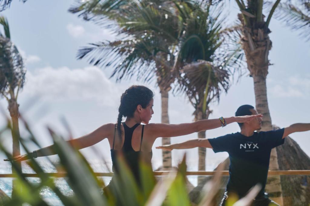 Divi cilvēki praktizē jogu pludmalē, apkārt palmām un saulainai debesskrāpju ainavai.