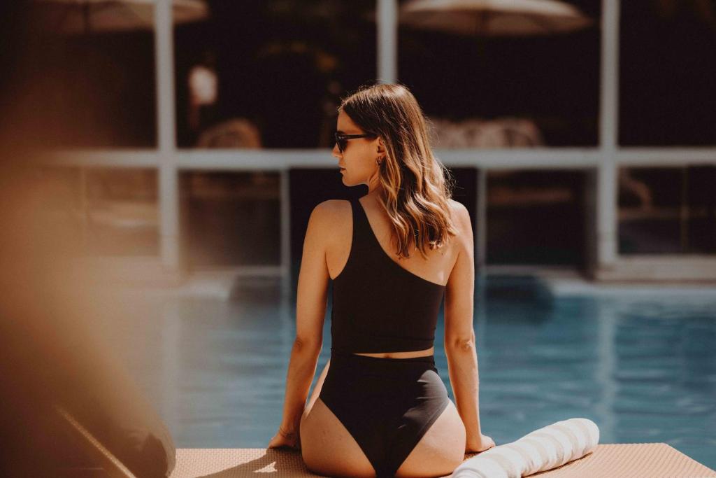 Jauna sieviete melnā peldkostīmā sēž uz saulessarga pie baseina, skatoties uz ūdeni. Apkārt ir relaksējoša atmosfēra ar pludmales krēsliem un saules stariem.