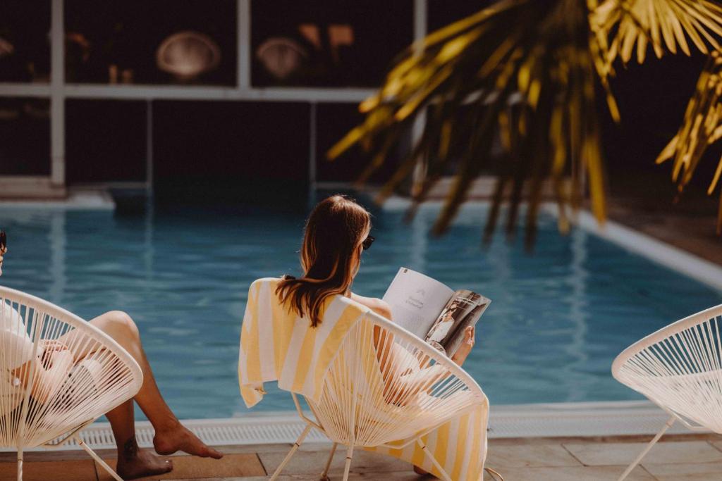 Sieviete atpūšas pie baseina, lasot žurnālu, tērpusies svītrainā peldkostīmā. Apkārt redzami baseins un koki, kas rada relaksējošu atmosfēru.