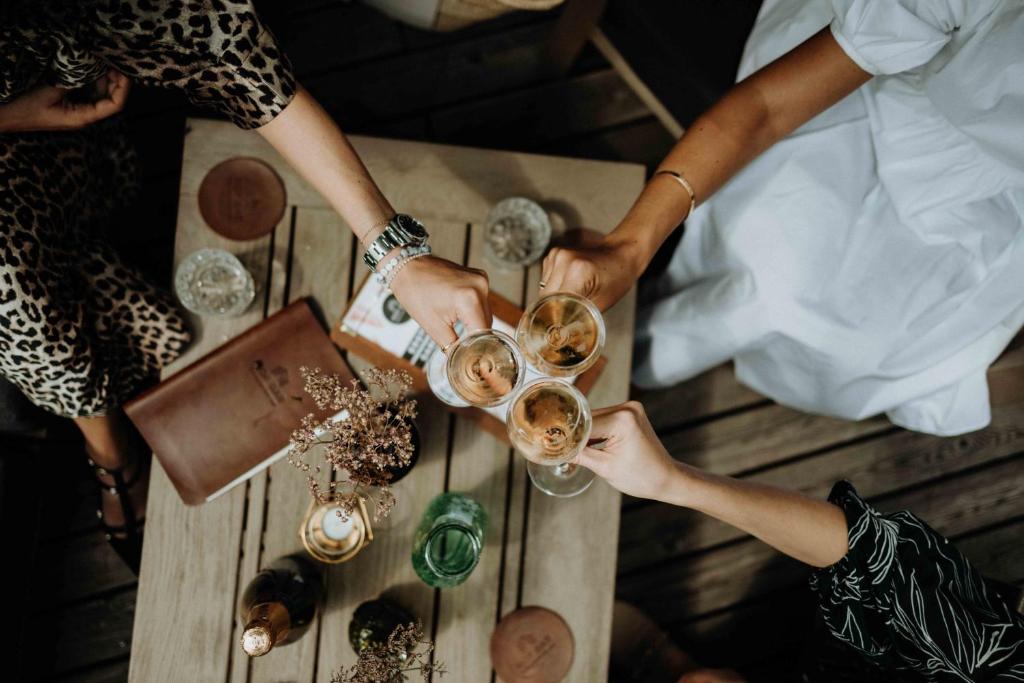 Sievietes pacelt glāzes, svinot kopā pavadītu laiku, uz koka galda. Apkārt ir dzērieni un dekoratīvi elementi, radot jautru un relaksējošu atmosfēru.