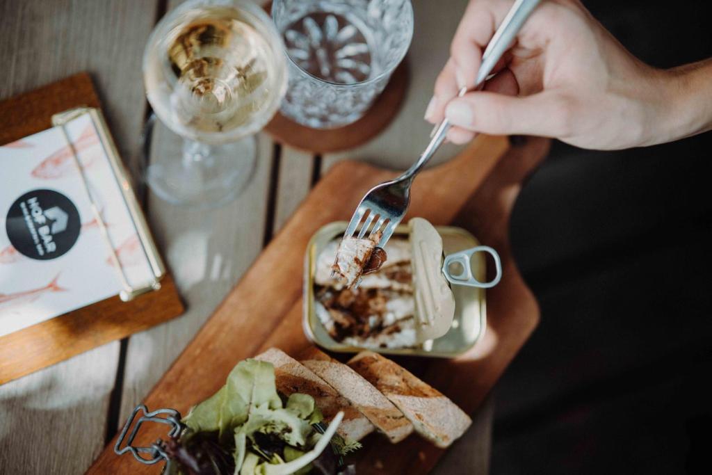 Gourmet ēdiens uz koka dēļa, kurā redzama atvērta konservu bundža ar zivīm, šķēles maizes un salāti, kopā ar glāzi baltvīna un ūdens glāzi.