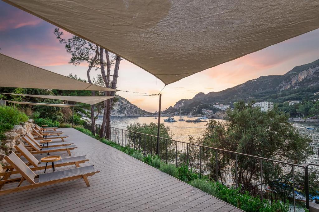 Relaksējoša ainava ar terasi, kurā ir sauļošanās krēsli un nojumes, kas piedāvā ēnu. Skats uz gleznaino ostu un kalniem saulrieta gaismā.