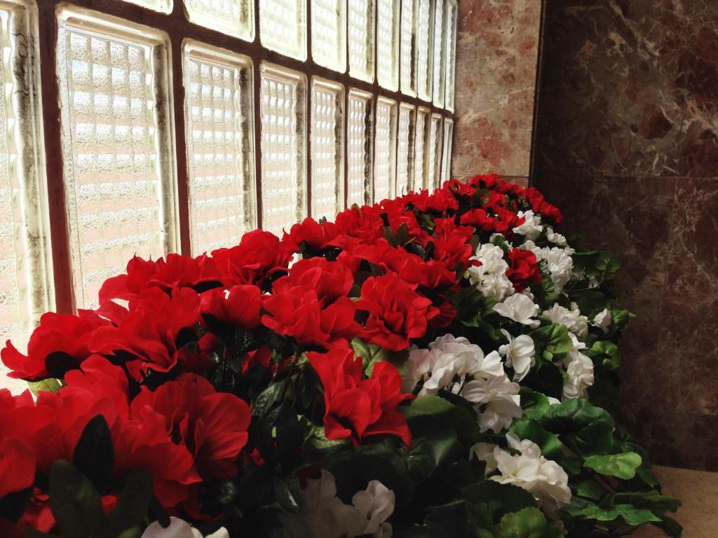 Sarkano un baltu mākslīgo ziedu kompozīcija, novietota pie stikla blokiem, kas ļauj cauri plūst gaismai. Ziedi ir sulīgi un dzīvīgi, radot siltu un viesmīlīgu atmosfēru.