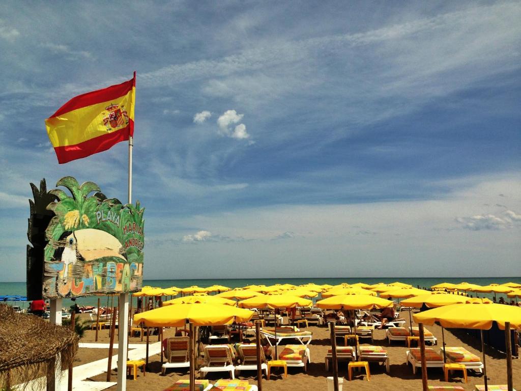 Pludmales ainava ar dzeltenām saulessargiem un Spānijas karogu, kas plīvo virs krasta. Attēlā redzama pludmales zona ar koka zīmi, kas norāda uz "Playa Marinas".