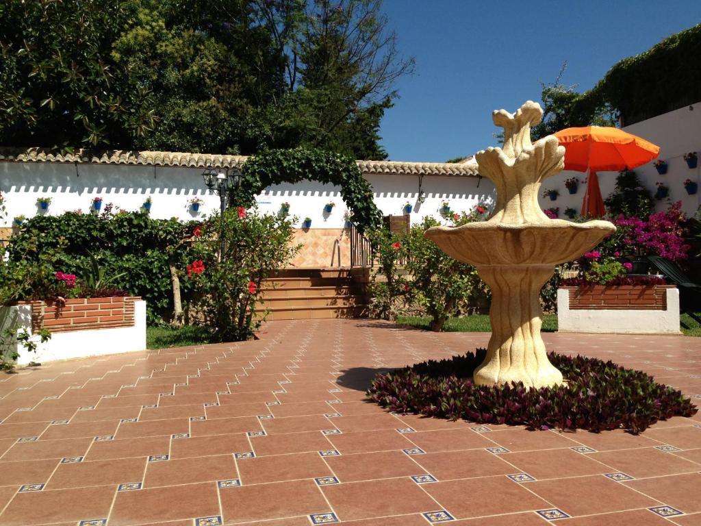 Dārzs ar stilīgu strūklaku, ko ieskauj ziedi un augi, saulainā dienā. Redzama oranža saulessarga un akmens flīžu grīda.